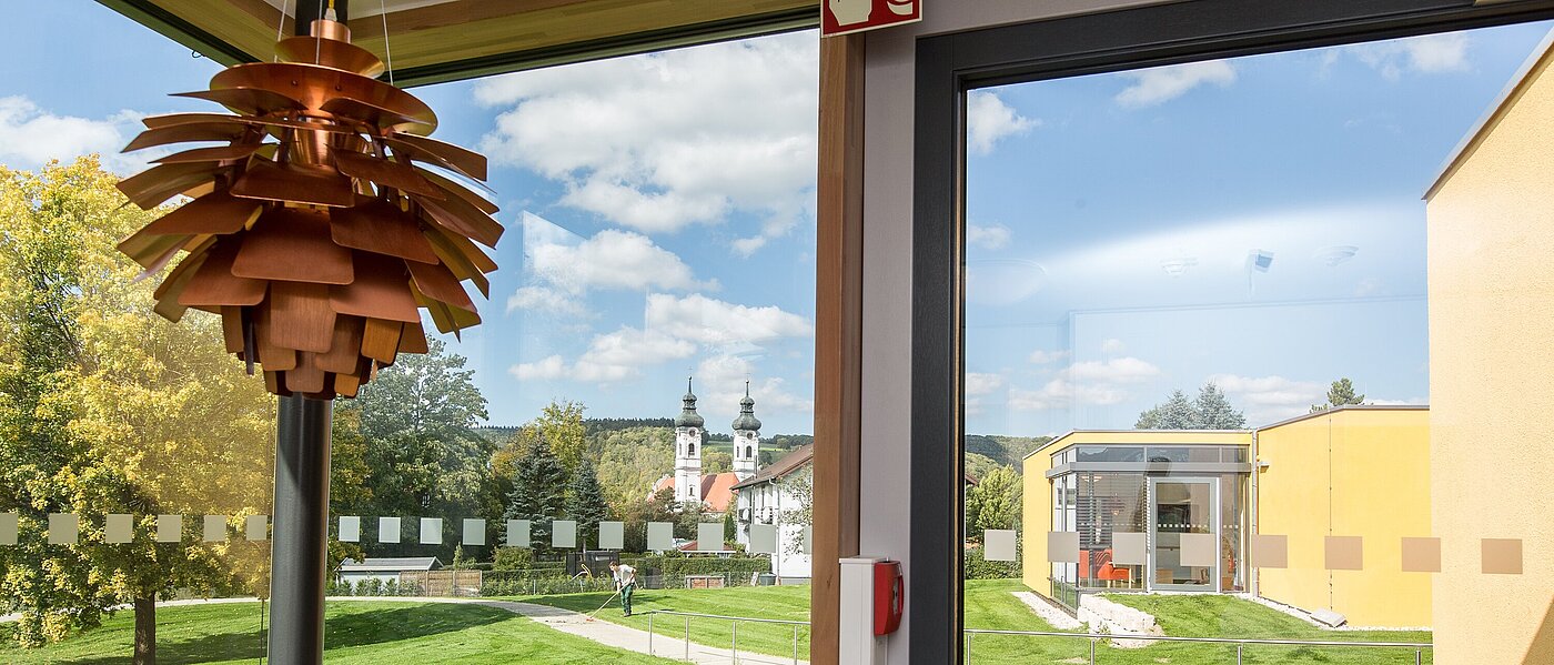 Man blickt aus einem großen bodentiefen Fenster ins Grüne, im Hintergrund sind die Türme des Zwiefalter Münster zu sehen. 