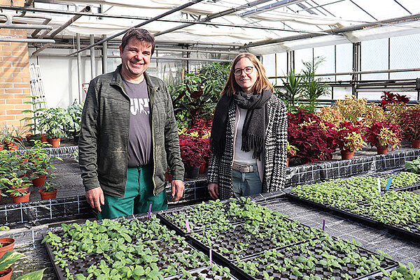 Ein Mann in Arbeitskleidung und eine junge Frau stehen lächelnd an langen Pflanztischen in einem Gewächshaus.
