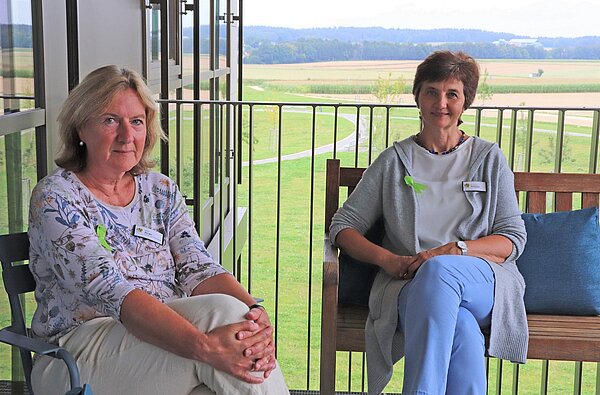 Zwei Frauen sitzen auf einem Stuhl und einer Bank auf einem Balkon. Hinter ihnen der Blick auf Felder und Wald.