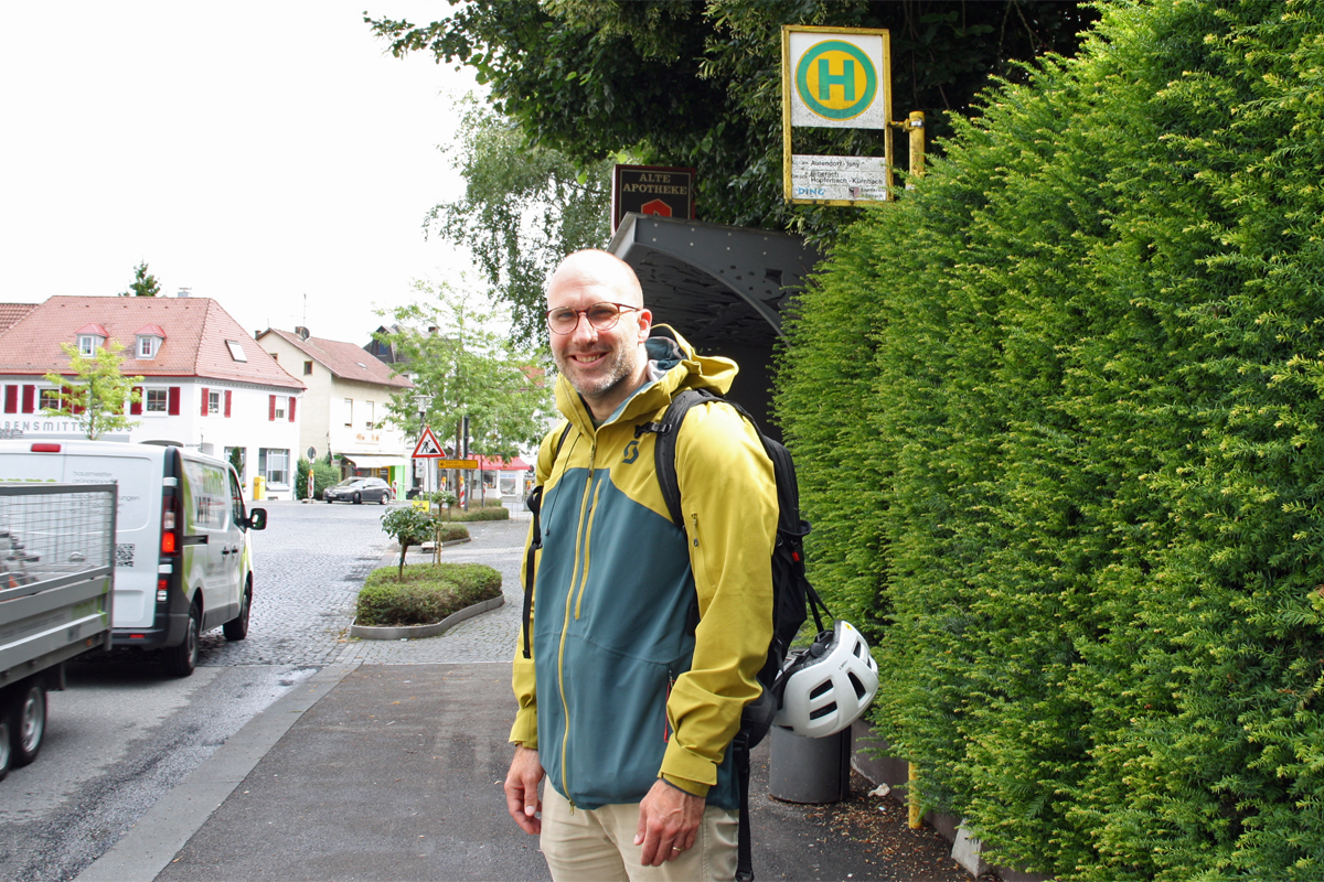 An einer Bushaltestelle an einer Straße steht ein Mann, der freundlich lächelt. Er trägt eine Brille, eine bunte Regenjacke und einen Rucksack, an dem ein Fahrradhelm hängt. Links im Bild fährt ein Transporter mit Anhänger vorbei.