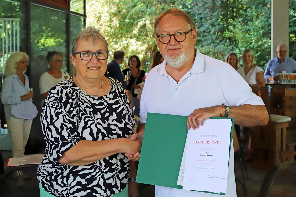 Eine Frau und ein Mann reichen sich die Hand und lächeln dabei in die Kamera, im Hintergrund sind die Gäste der kleinen Feierlichkeit im Freien zu erkennen.