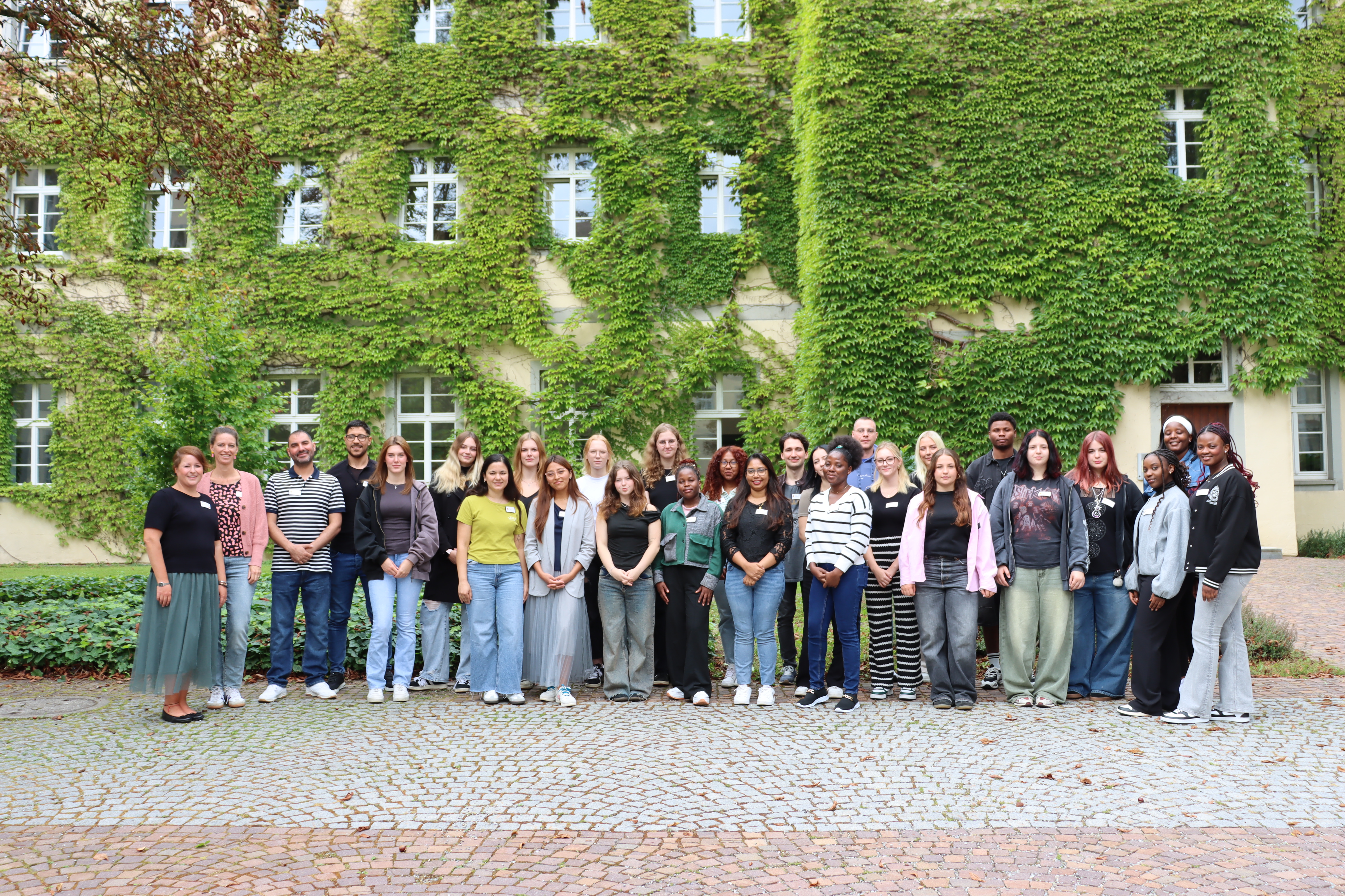 Eine große Gruppe jüngerer Menschen steht vor einer mit Efeu bewachsenen Hauswand.