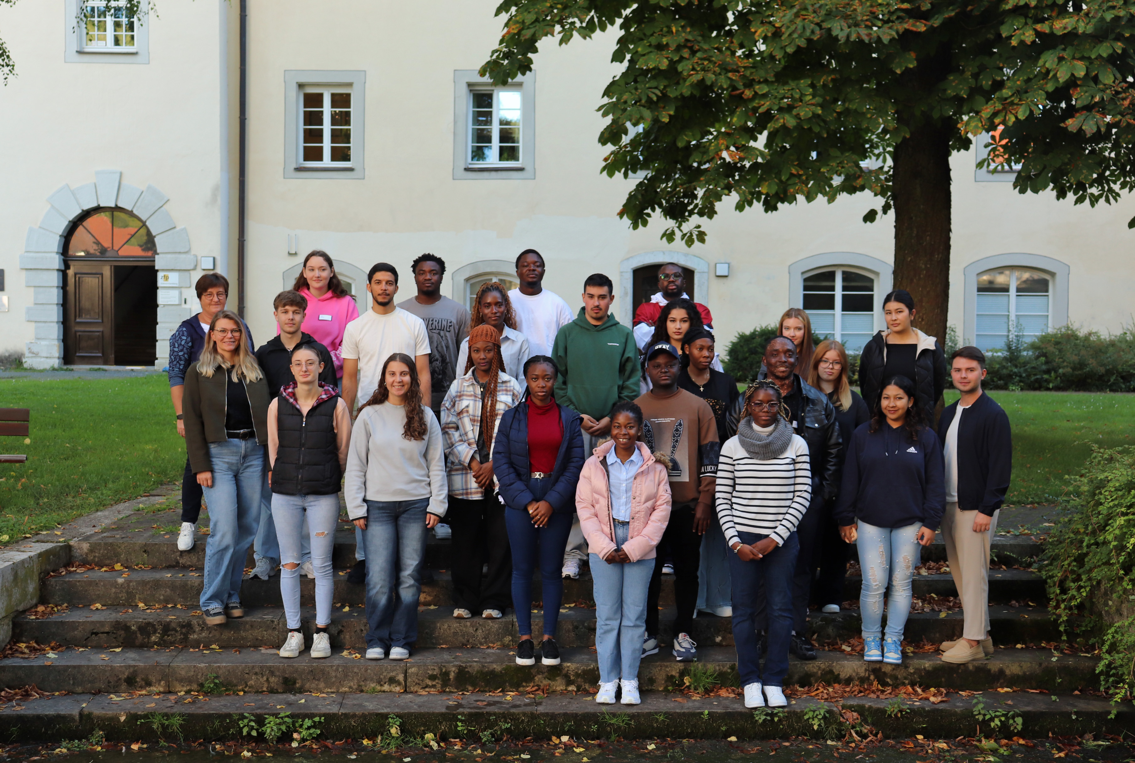 Gruppenfoto von 20 Personen, die auf einer Treppe vor einem Gebäude stehen. Die meisten tragen Freizeitkleidung, einige mit Jacken und Pullovern. Bäume im Hintergrund.