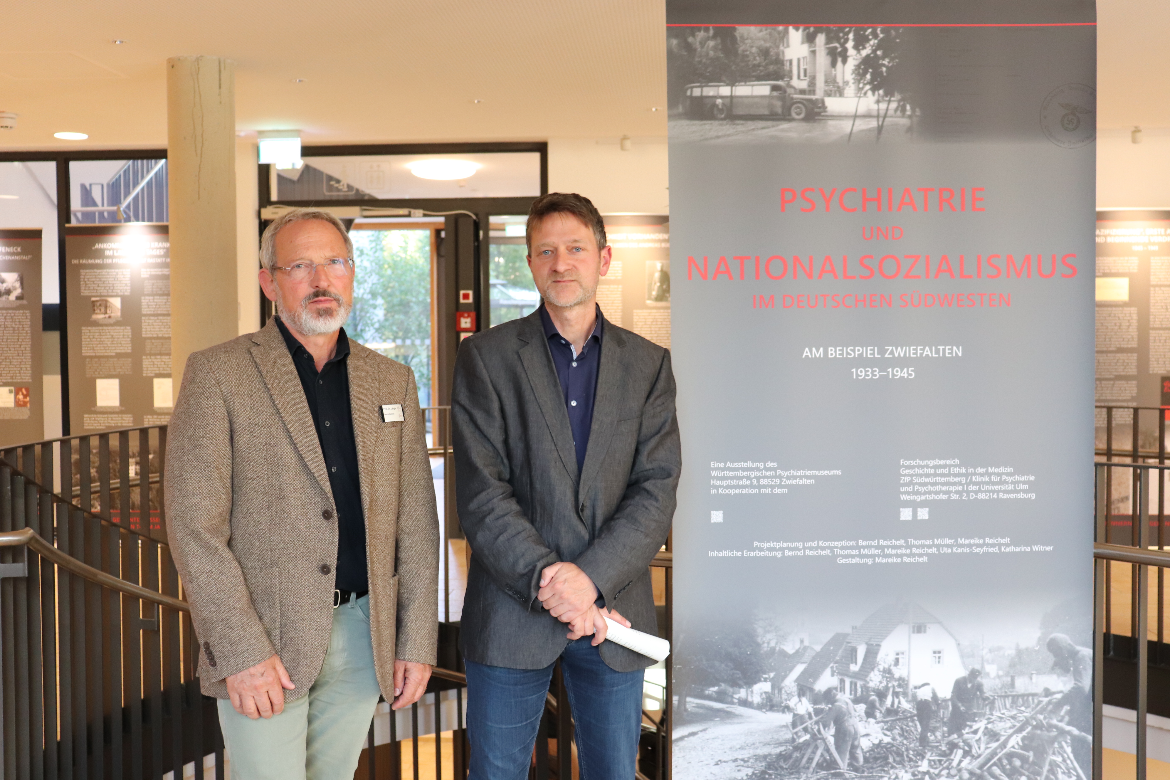 Zwei Personen stehen neben einem Informationsbanner zur Wanderausstellung 'Psychiatrie und Nationalsozialismus im deutschen Südwesten 1933-1945'. Das Banner zeigt historische Fotos und Text.