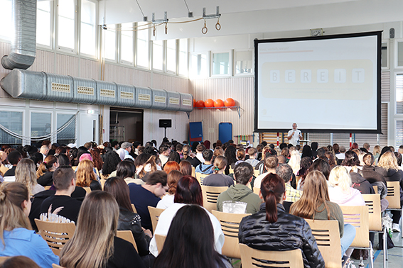 In einer Sporthalle sind viele Stühle aufgestellt, die Halle ist voll besetzt. Der Blick geht vom hinteren Ende über die Köpfe hinweg nach vorne, wo ein Mann am Rednerpult steht.